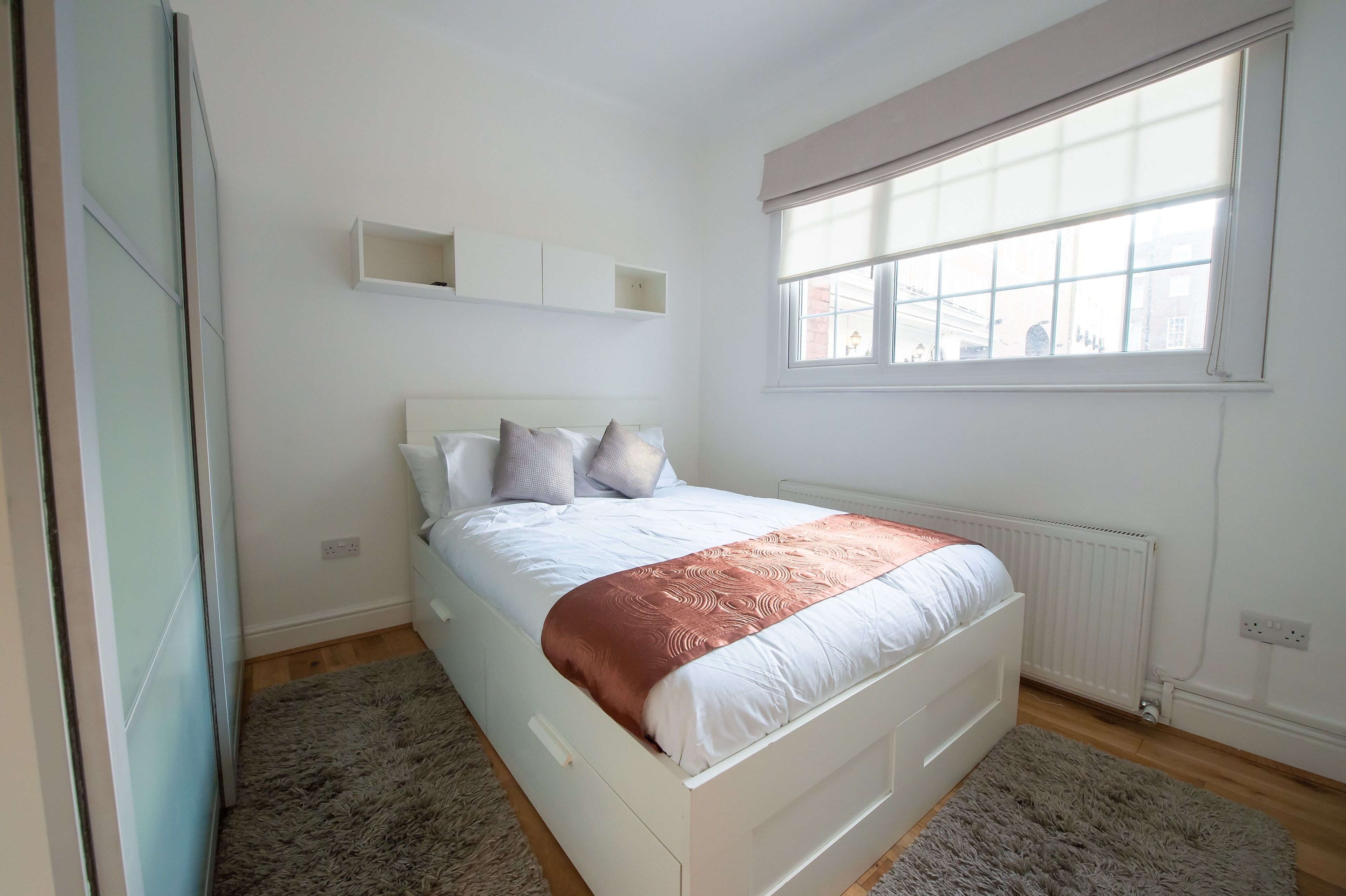 A bedroom with a bed, a window, a radiator, and a wall-mounted shelf.