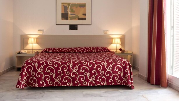 A hotel room with a bed, two bedside tables, a nightstand, a framed picture on the wall, and a window with curtains.