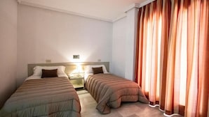 Una habitación de hotel con dos camas individuales, una mesita de noche con lámpara y un ventanal con cortinas.