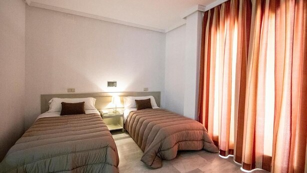 A hotel room with two single beds, a nightstand with a lamp, and a large window with curtains.