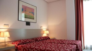 A hotel room with two beds, each with a red patterned bedspread, bedside lamps, a framed picture on the wall, and a window with curtains.