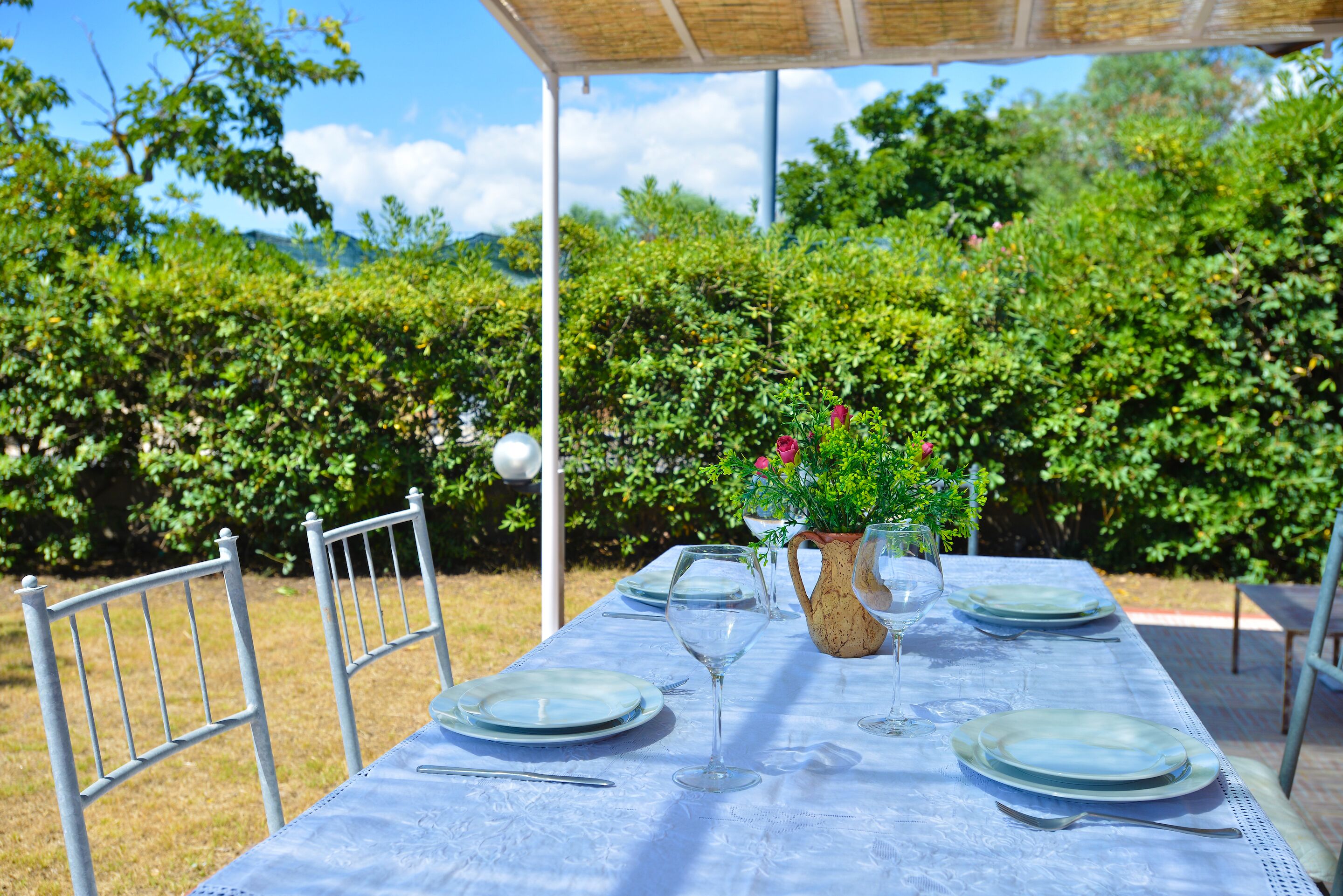A table set outdoors with a blue tablecloth, metal chairs, and a vase with flowers.