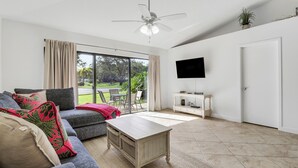 A spacious living room with a grey sofa, a wooden coffee table, a ceiling fan, and a TV mounted on the wall.