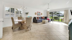 A spacious living area with a dining table and chairs, a sofa, and a large window with blinds.