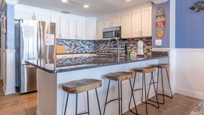 A kitchen with white cabinets, a stainless steel refrigerator, a microwave, and a tiled backsplash. There are four wooden stools at the island.