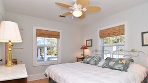 A bedroom with a bed, two windows with blinds, a ceiling fan, a bedside table with a lamp, and a framed picture on the wall.