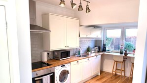 A modern kitchen with white cabinets, a built-in oven, microwave, and washing machine. There are wooden countertops, a sink, and a window with a view of the outside.