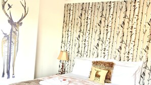 A neatly made bed with a patterned bedspread, a wall with a birch tree wallpaper, and a wall-mounted lamp.