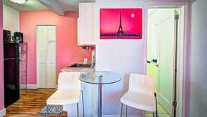 A compact kitchen with a glass table and two white chairs, a refrigerator, and a coffee maker.