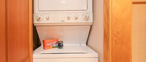 A compact laundry room with a stacked washer and dryer, a box of Bounce detergent, and a jar of detergent.