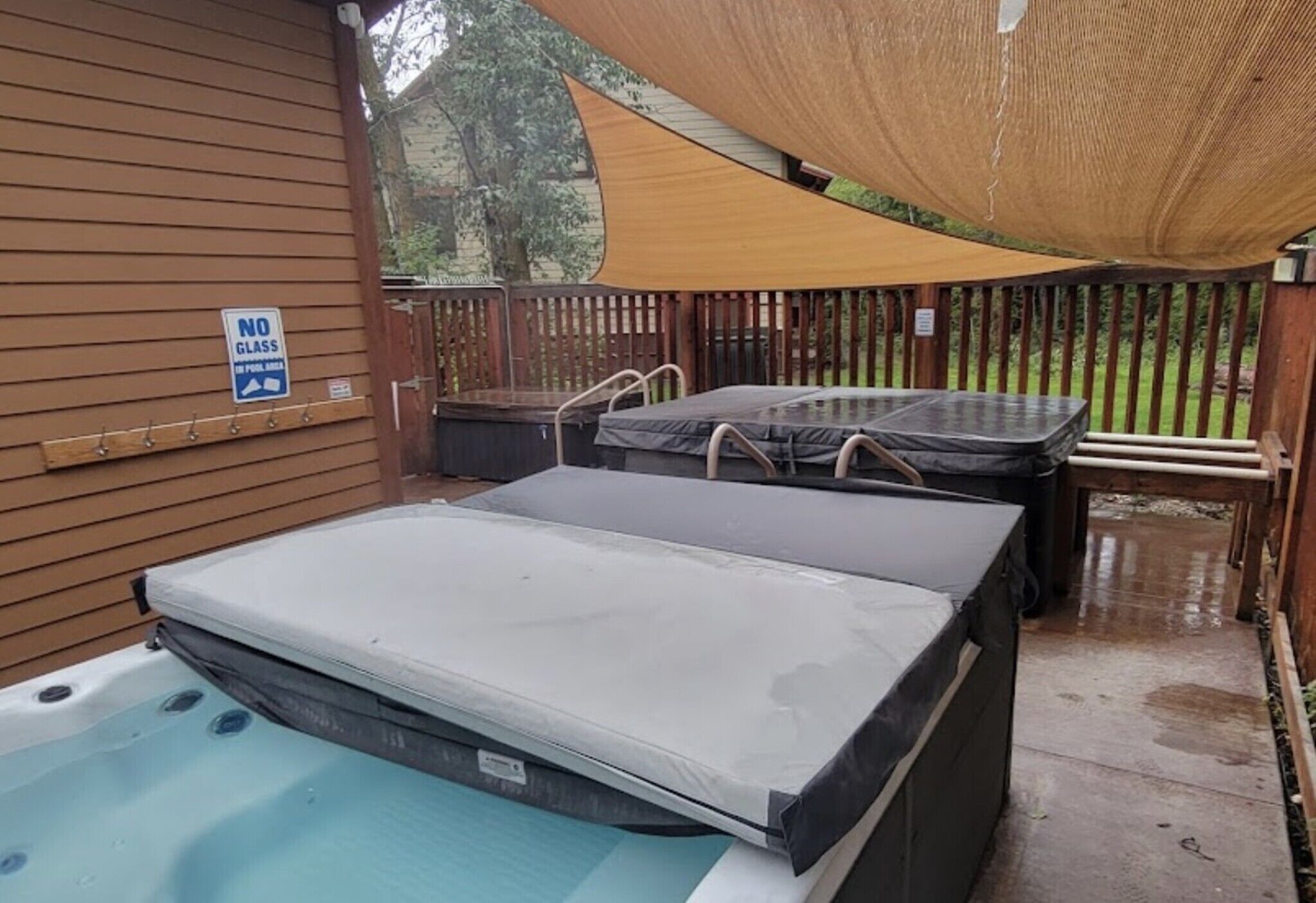 Un jacuzzi con tapa, una terraza de madera, un cartel de prohibición de vidrios y una zona con sombra.