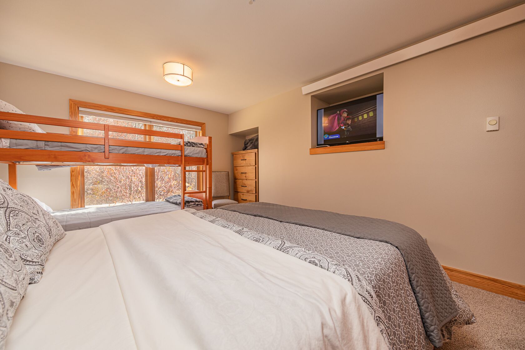 Quarto com beliches, televisão presa na parede e janela com vista para árvores.