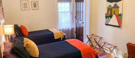 A room with two beds, a small table, a chair, a lamp, and a painting on the wall.
