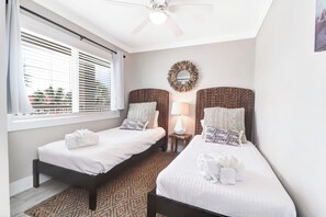Ein Schlafzimmer mit zwei Betten, einem Deckenventilator, einem Fenster mit Jalousien, einem Wandspiegel und einem kleinen Tisch mit Lampe.