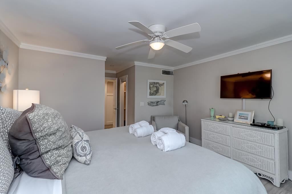Ein Hotelzimmer mit einem Bett, einem Deckenventilator, einem Fernseher, einem Schminktisch und einer Tür zu einem anderen Raum.