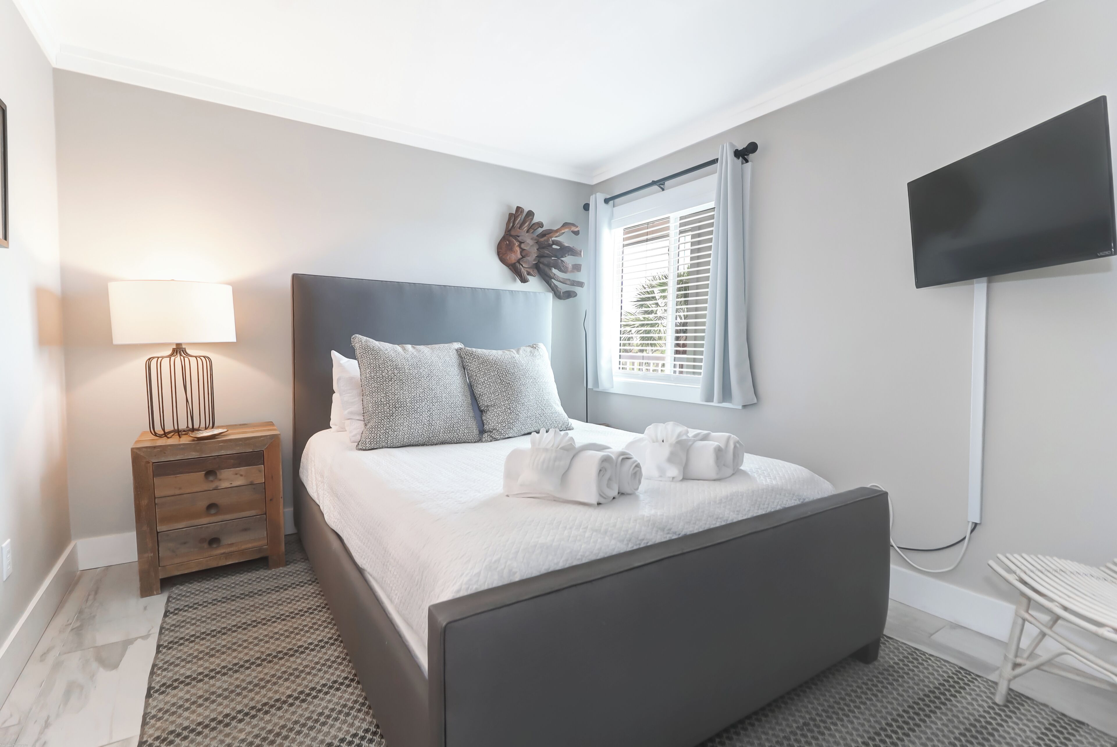 Une chambre à coucher moderne, avec un lit, une table de chevet, une lampe, un téléviseur fixé au mur et une fenêtre avec des stores.