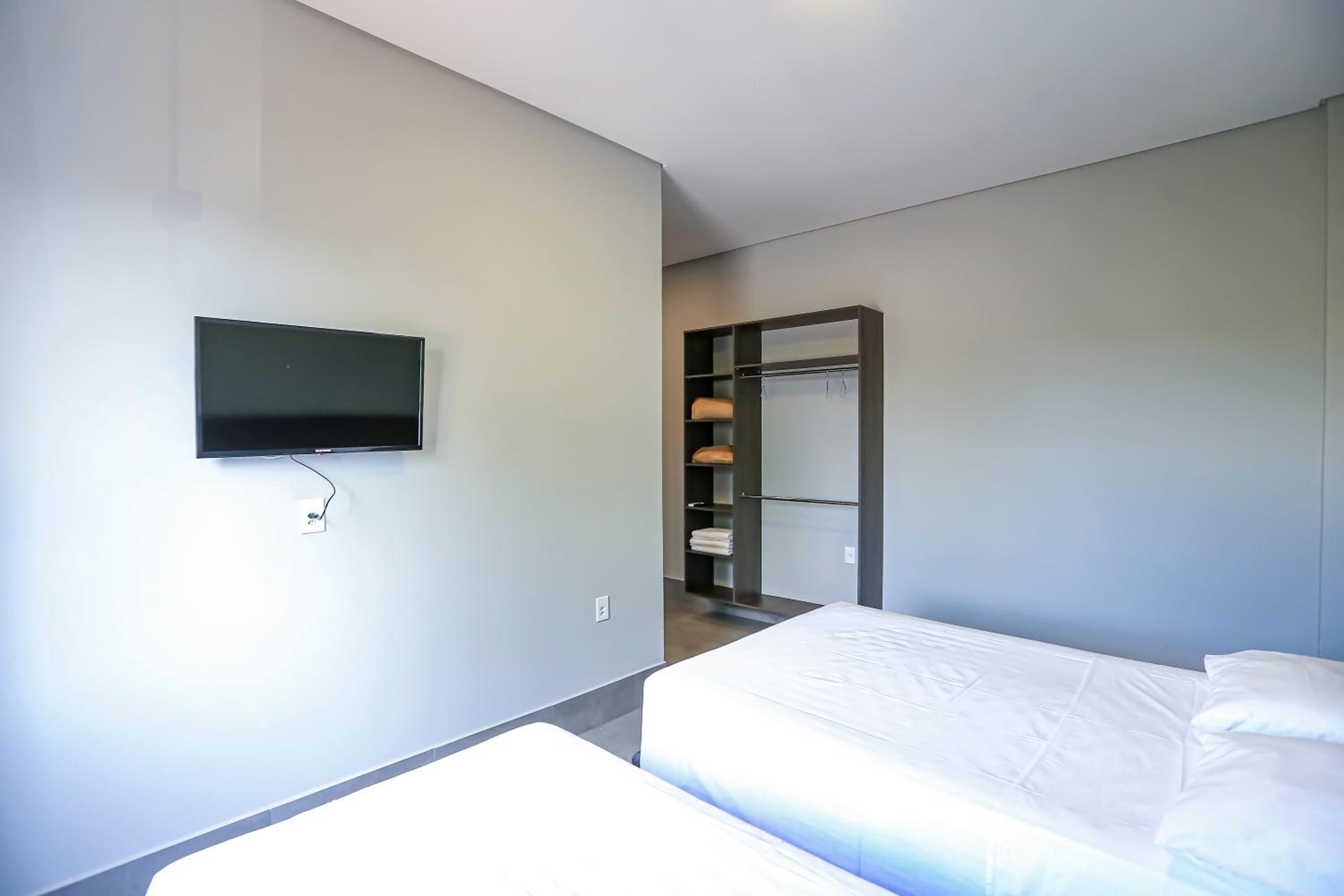 A modern hotel room with a bed, a TV mounted on the wall, and a walk-in closet with shelves and hanging space.