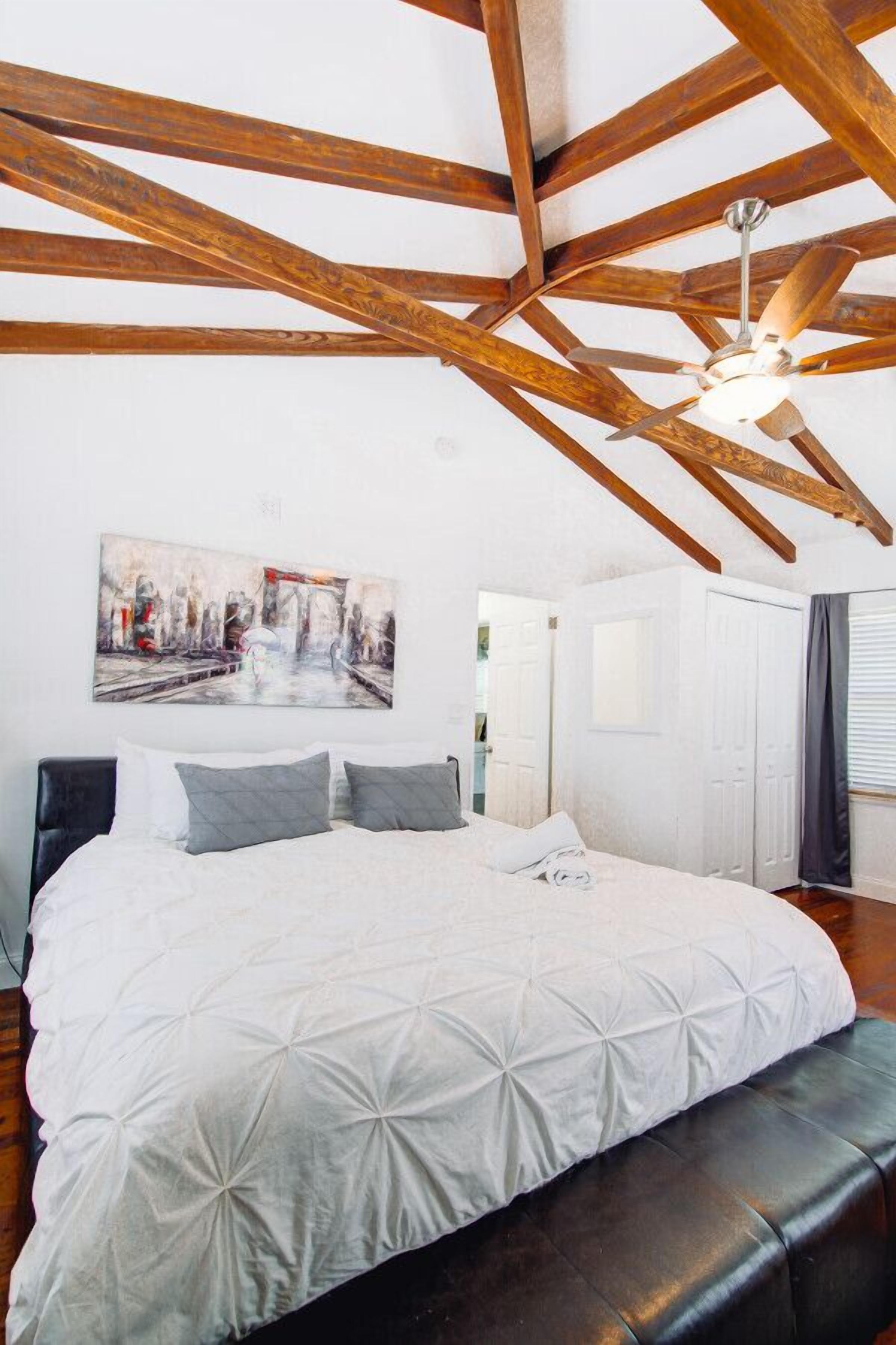 Een slaapkamer met een groot bed, houten balken, een plafondventilator en een schilderij van een stadsgezicht.