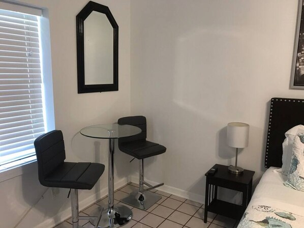 A small, modern room with a round glass table, two black chairs, a single bedside table with a lamp, a bed with a headboard, and a large mirror on the wall.