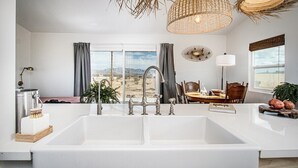 A modern kitchen with a large white sink, a dining area with a table and chairs, and a view of the desert landscape through the window.