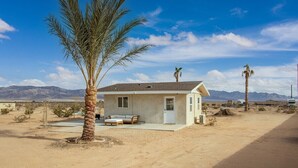 A small, single-story house with a front porch, a bed, and a door, surrounded by desert landscape and mountains.