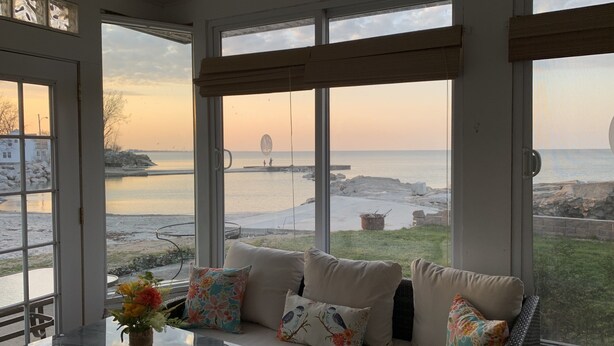 Una habitación iluminada por el sol con un amplio ventanal con vistas a la playa, un rincón de descanso con cojines y una mesa con un jarrón de flores.