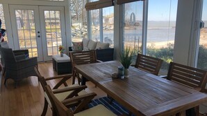 Un comedor amplio con una mesa y sillas de madera, un sofá y vista a la playa a través de ventanas grandes.