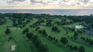 Un campo de golf con un cuerpo de agua al fondo, rodeado de árboles y edificios.