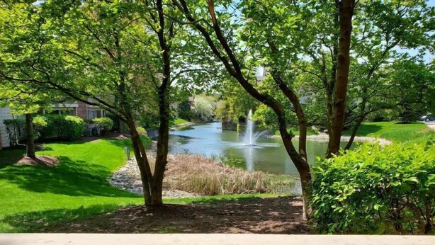 A view from a wooden deck overlooking a pond with a fountain, surrounded by lush greenery and a well-maintained lawn. - Cozy Condo - Pond View - Monthly Discounts- Washer & Dryer-Great Location (Lombard)