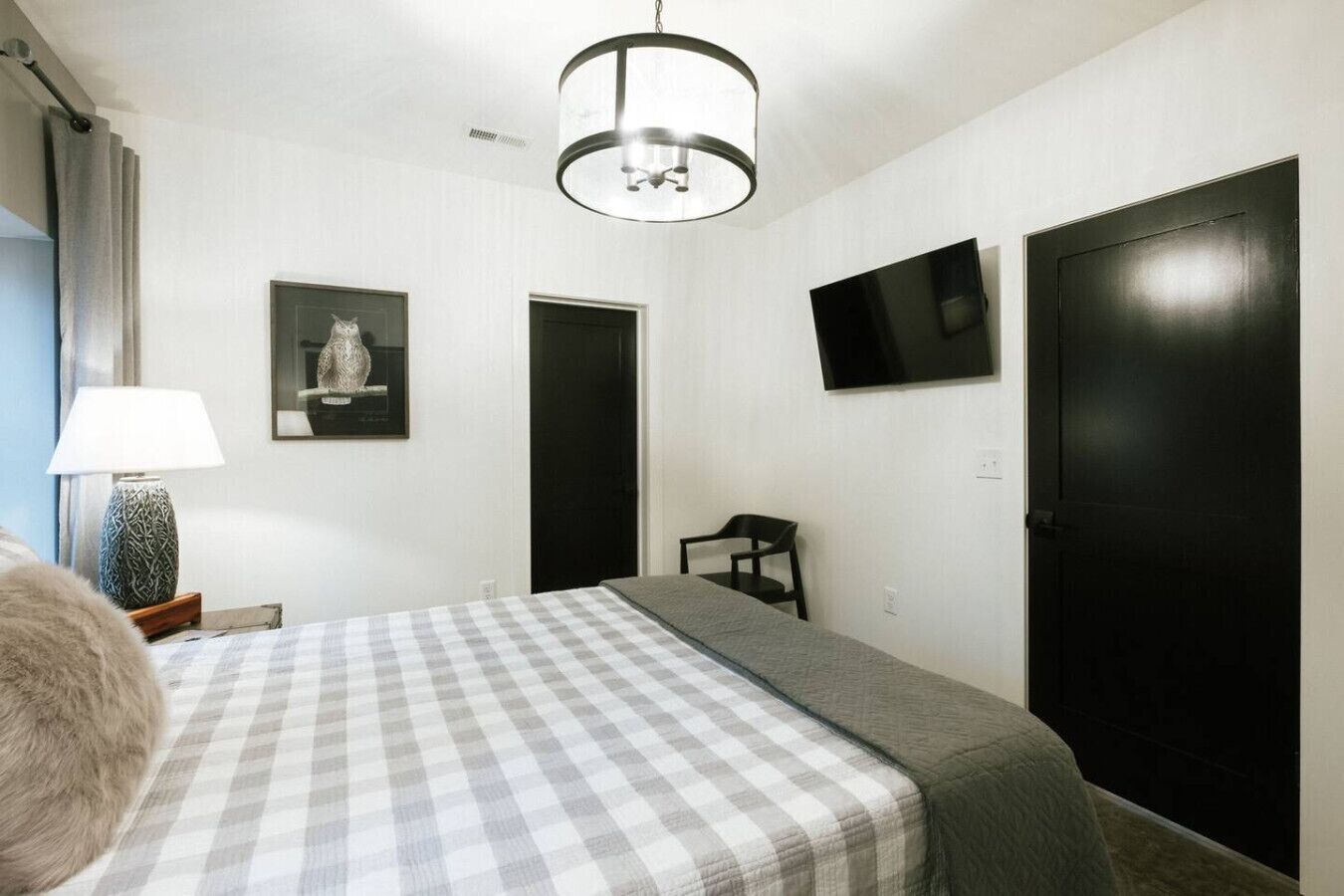A bedroom with a checkered bedspread, a bedside lamp, a framed picture, a television, and a black door.