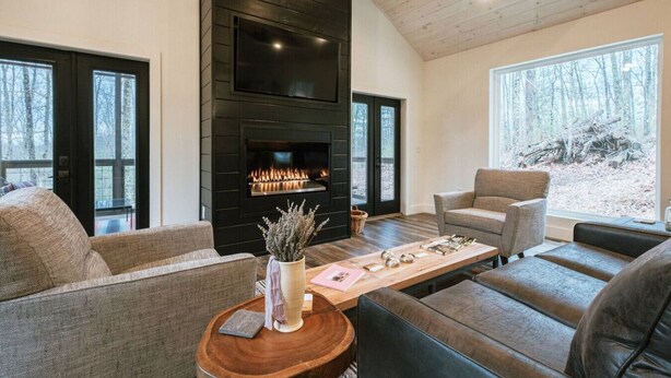 A cozy living room with a fireplace, two sofas, a coffee table, and large windows offering a view of the forest. - Honeysuckle Bridge- Hot Tub, Firepit, Private Hiking, Sandstone Arch (Campton)