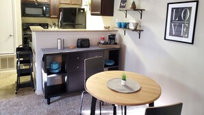 Uma cozinha compacta com eletrodomésticos modernos, uma área de refeições com mesa redonda e uma prateleira fixada na parede com objetos decorativos.