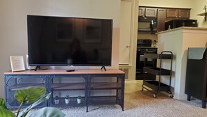 Uma sala de estar com televisão de tela plana em um móvel, uma mesinha, uma planta e uma área de cozinha com armários e eletrodomésticos.
