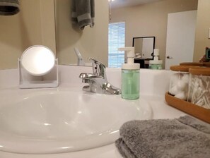 A bathroom with a white sink, a round mirror, a faucet, and a bottle of liquid soap. There are towels hanging on the wall and a window with blinds.