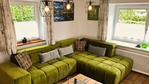 Un sofá seccional verde con múltiples cojines, una mesa de centro de madera con un vaso y un pequeño recipiente, y una ventana con cortinas y vistas a la vegetación.