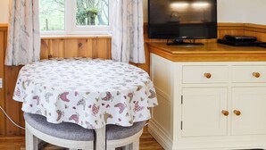 Une pièce avec une table ronde recouverte d’une nappe à motifs floraux, un téléviseur posé sur un meuble en bois et une fenêtre avec des rideaux.