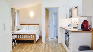 Une chambre compacte avec un lit en bois, une table de chevet et une fenêtre avec des rideaux. À côté se trouve une petite cuisine équipée d’un four à micro-ondes, d’un grille-pain, d’une bouilloire et de placards.