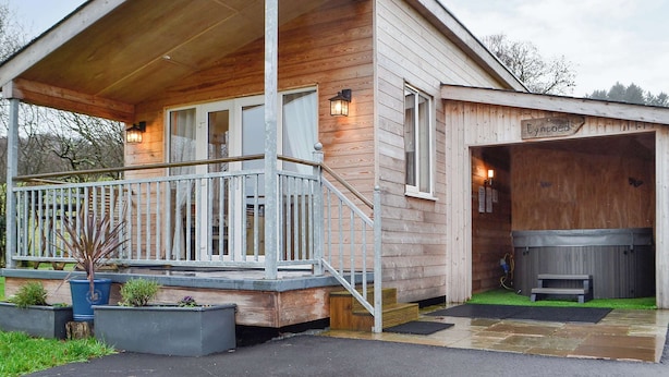 Une cabane en bois dotée d’un porche couvert, d’un jacuzzi et d’une enseigne indiquant « Eyncoed ».