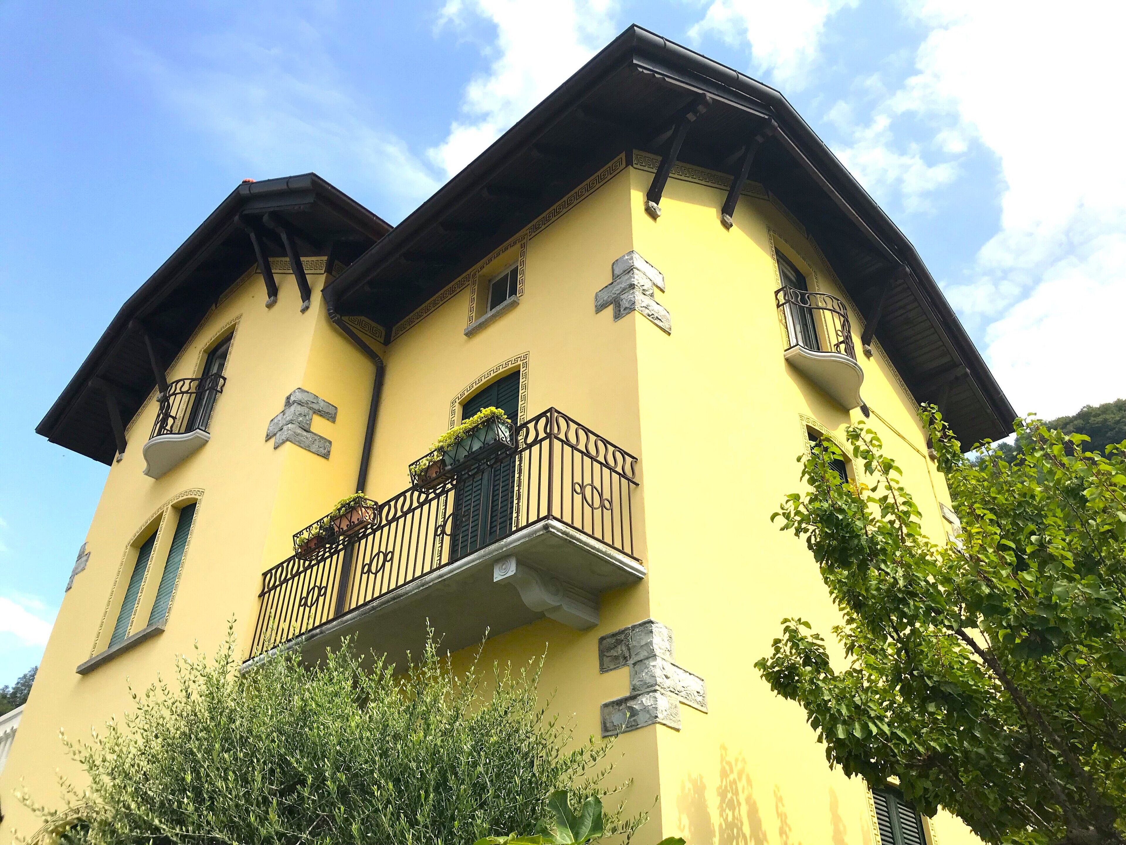 Un edificio amarillo con balcón, pared de piedra y ventana con contraventanas.