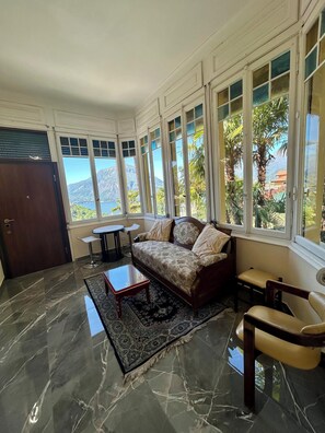 Ein Zimmer mit einer Couch, einem kleinen Tisch, einem Sessel und Blick auf Berge und Grün durch große Fenster.