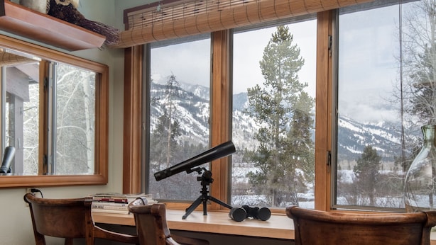 Une pièce comprenant un bureau, deux chaises, un télescope et une grande fenêtre donnant sur des arbres et des montagnes.