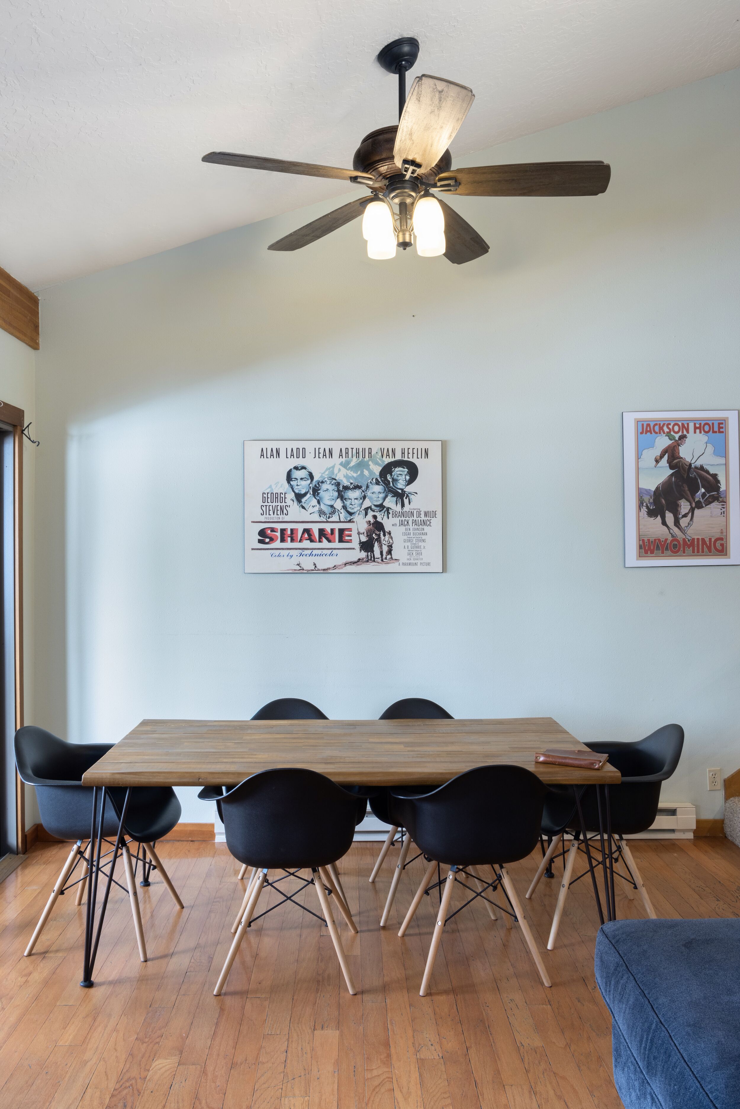 Un coin repas avec une table en bois et des chaises noires, un ventilateur de plafond et deux affiches accrochées au mur.