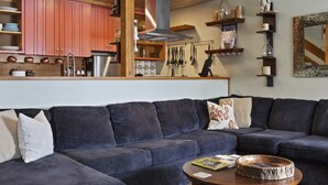 Un salon avec un canapé d’angle bleu, une table basse ronde et un tapis en peau de vache. On aperçoit une cuisine à l’arrière-plan, avec des meubles en bois et un luminaire suspendu.