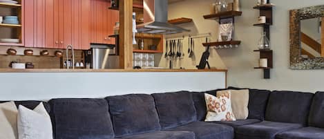 A living room with a blue sectional sofa, a round coffee table, and a cowhide rug. There is a kitchen visible in the background with wooden cabinets and a hanging light fixture.