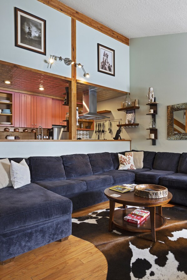 A living room with a blue sectional sofa, a round coffee table, and a cowhide rug. There is a kitchen visible in the background with wooden cabinets and a hanging light fixture. - Outpost: Twinberry 3522 - Located in the Aspens! (Wilson)