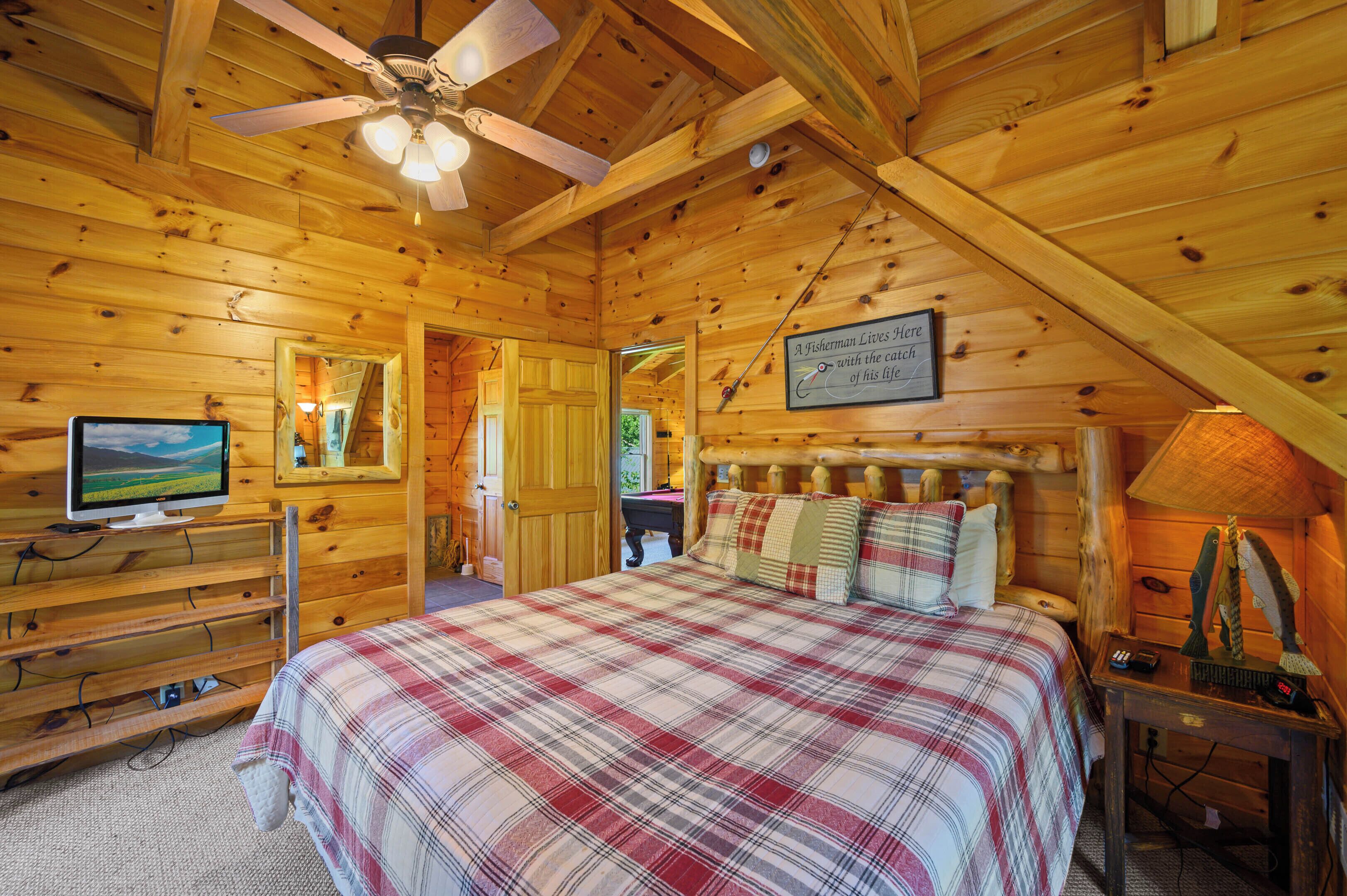 A wooden cabin bedroom with a bed, a television, and a ceiling fan.