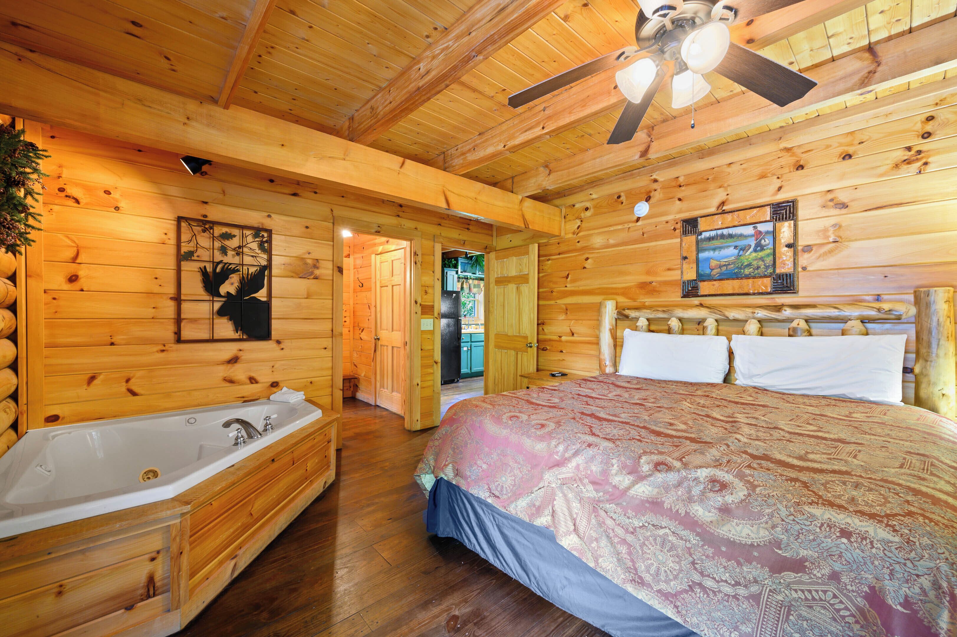 A log cabin bedroom with a large bed, a wooden headboard, a ceiling fan, and a bathroom with a bathtub.