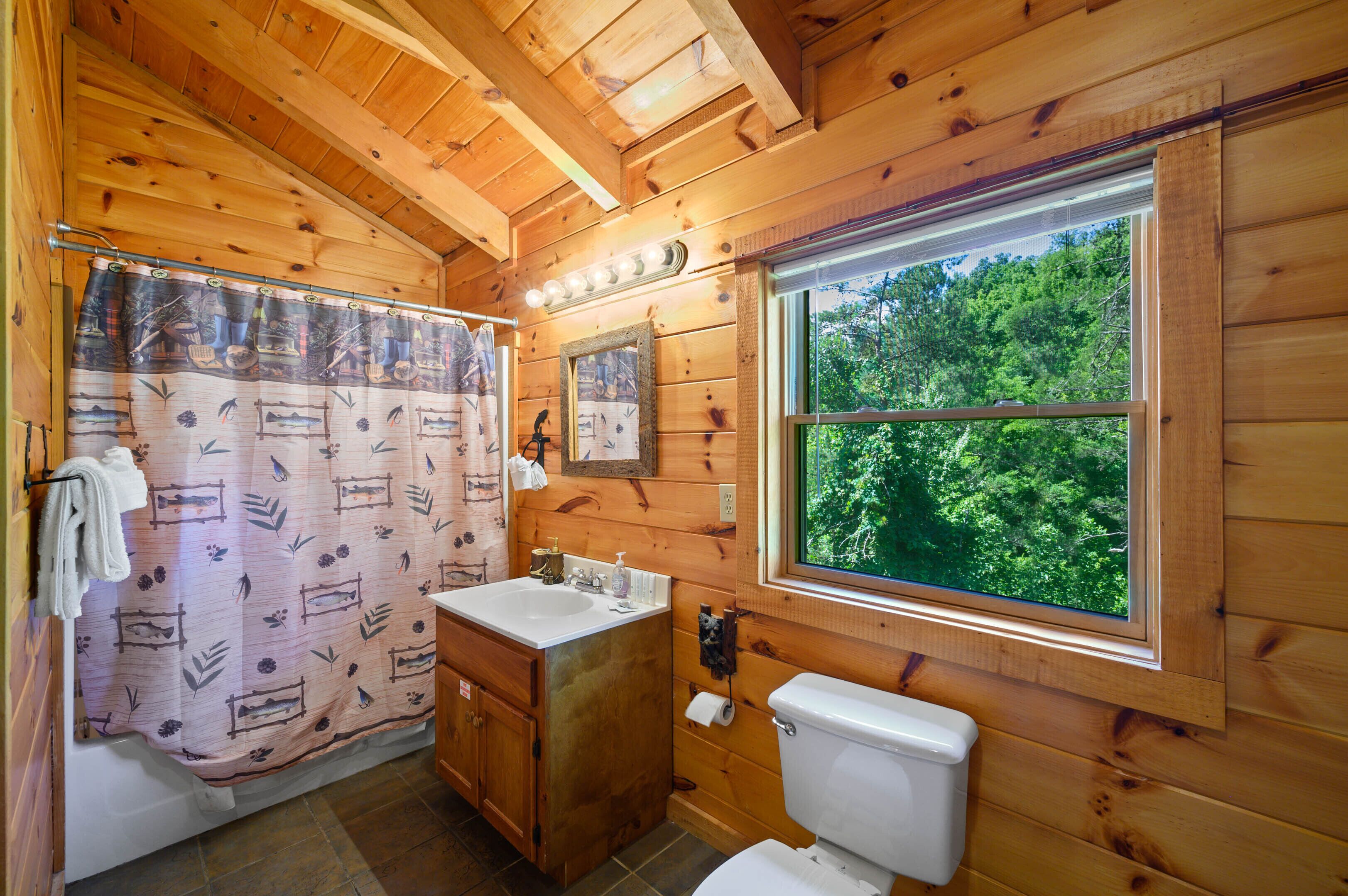 Baño de cabaña de madera con cortina de ducha, lavamanos, inodoro y ventana con vistas al bosque.