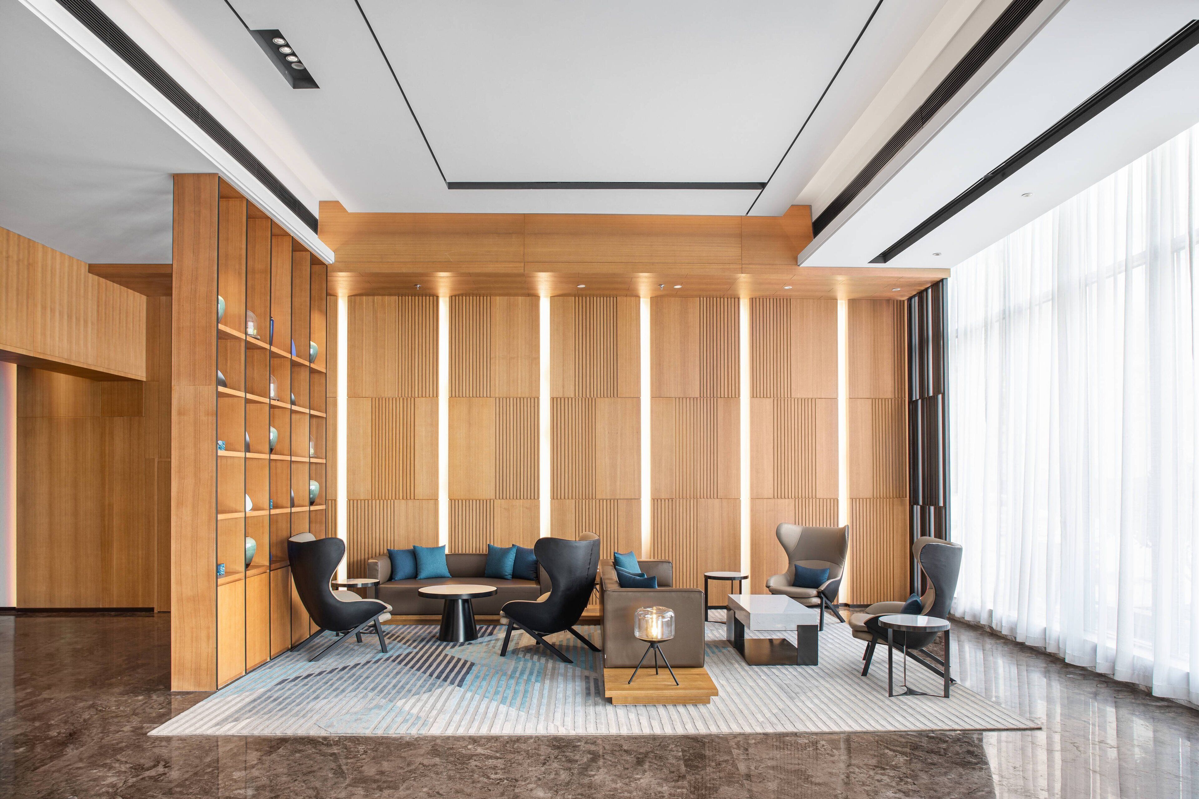 A modern lobby with a wooden panel wall, a seating area with sofas and chairs, and a large window providing natural light.
