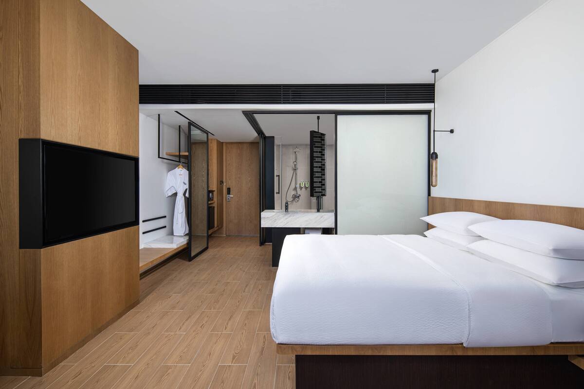 Una habitación de hotel moderna con una cama grande, un armario de madera, televisión integrada y un baño con ducha y lavabo.
